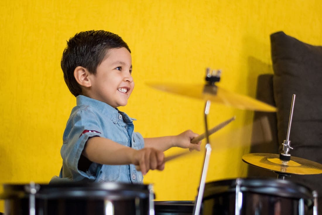 A young boy grinning behind a drum kit, sticks mid-swing